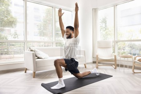 Feliz positivo deportivo africano chico entrenamiento cuerpo en yoga mat, haciendo embestidas con las manos levantadas, mirando hacia otro lado, sonriendo, disfrutando de ocio activo, aptitud en el hogar, estirando los músculos de las piernas