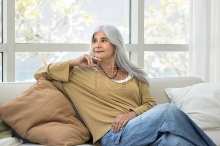Foto de Mujer mayor soñadora positiva relajándose en un cómodo sofá en una acogedora casa, mirando hacia otro lado, tocando la barbilla, pensando, soñando despierto con una sonrisa reflexiva, disfrutando de la jubilación, el ocio - Imagen libre de derechos