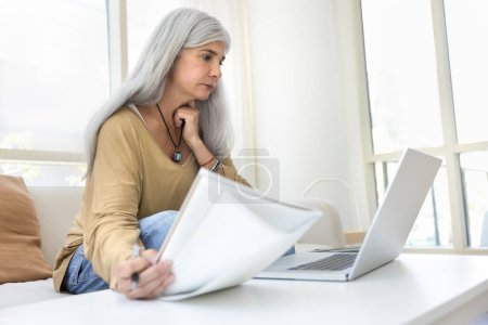 Foto de Enfocado mayor mayor madura latina mujer estudiando en línea en casa, trabajando en el ordenador portátil, sosteniendo portátil para escribir, viendo el aprendizaje de contenido de vídeo en Internet - Imagen libre de derechos