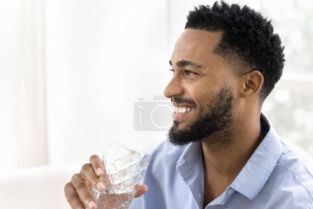 Foto de Alegre guapo afroamericano bebiendo agua clara, sosteniendo el vaso con bebida fresca, cuidando un estilo de vida saludable, nutrición, equilibrio acuático, hidratación, mirando hacia otro lado, sonriendo - Imagen libre de derechos