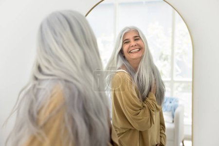 Photo for Cheerful carefree attractive senior Latin woman with long grey hair looking in mirror, smiling, laughing, enjoying reflection, wellness, dancing, posing with flying hair - Royalty Free Image