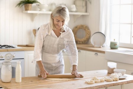 Photo for Beautiful middle-aged housewife cooking in modern cozy kitchen. Smiling mature woman in apron flattening dough with rolling-pin, baking sweet buns alone at home. Culinary, homemade food preparation - Royalty Free Image