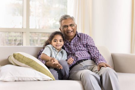 Téléchargez les photos : Heureux grand-père indien plus âgé et mignon petit-fille enfant étreignant avec amour, affection, assis sur le canapé de la maison, regardant la caméra, souriant pour le portrait de famille, riant - en image libre de droit