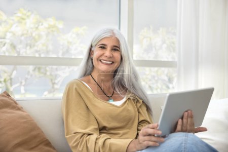 Photo for Happy senior student woman enjoying online learning application on tablet. Cheerful mature grey haired Latin lady holding tablet, looking at camera, smiling, laughing, posing for home portrait. - Royalty Free Image