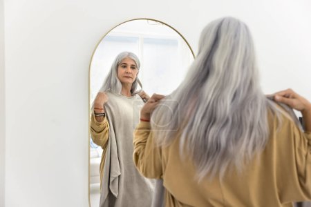 Téléchargez les photos : Femme hispanique âgée mature sérieuse essayant un pull surdimensionné, regardant dans le miroir, prenant soin de la mode, du style, de la tenue, choisissant un tissu tendance à porter, vérifiant l'achat sur la boutique en ligne - en image libre de droit