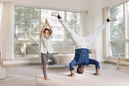 Téléchargez les photos : Joyeux père yogi indien et petite fille enfant faisant du yoga à la maison, gardant la tête et la pose de l'arbre, regardant la caméra, souriant, faisant de l'exercice ensemble, appréciant l'activité - en image libre de droit