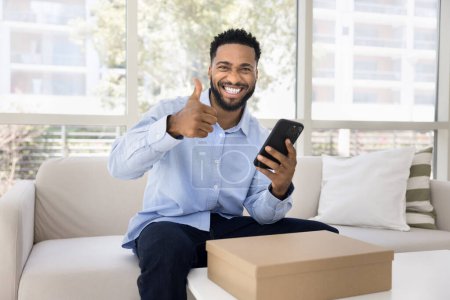 Photo for Cheerful satisfied young African client man making like hand gesture, showing thumb up at camera, smiling for portrait, using transportation online service over logistic cardboard box - Royalty Free Image