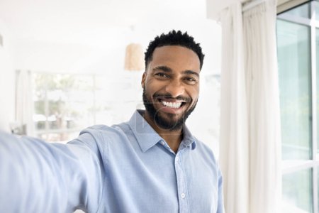 Photo for Cheerful handsome young African man taking selfie, looking at camera with toothy smile, showing perfect white teeth, holding gadget with webcam in outstretched hand, talking on video call - Royalty Free Image