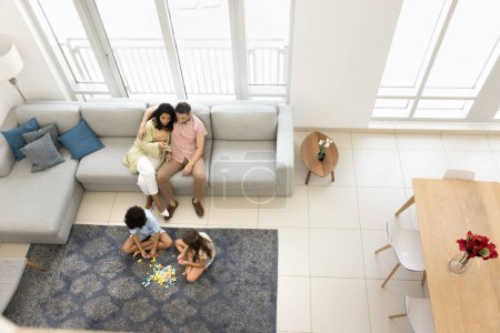 Photo for Couple of young parents watching two little sibling kids playing on heating carpeted floor, resting on home couch, hugging, relaxing in comfortable apartment interior. Top view, wide shot - Royalty Free Image