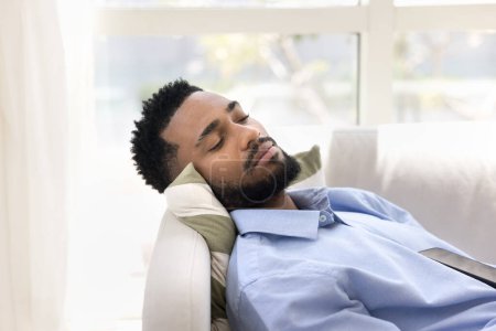 Téléchargez les photos : Sérénité somnolente attrayant jeune homme d'affaires africain se détendre pendant la pause de travail, dormir sur le canapé pendant la journée, faire la sieste, profiter de la paix, détente, loisirs, se sentir fatigué, épuisé - en image libre de droit