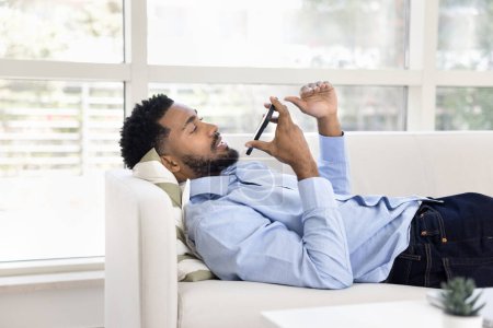 Photo for Serious young African business man talking on speaker on mobile phone, resting on couch in office hall, speaking at smartphone, recording voice audio message on work break - Royalty Free Image