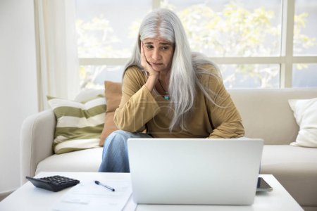 Sad unhappy retired elderly woman looking at accounting work result on laptop screen, touching chin, thinking on financial problems, overspending, high expenses, bankruptcy risk