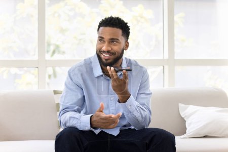 Photo for Positive handsome young African business man talking on speaker on cellphone, recording voice message, looking away, thinking, activating modern smart home application - Royalty Free Image