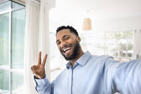Photo for Happy joyful African American guy showing victory winner fingers sign and toothy smile, looking at camera, making peace hand gesture, taking selfie portrait, enjoying success, laughing - Royalty Free Image
