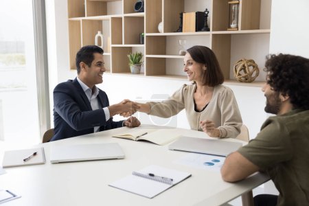 Positive successful multiethnic business partners shaking hands over meeting negotiations table, talking, smiling, laughing, getting agreement, ending briefing with handshake greeting gesture