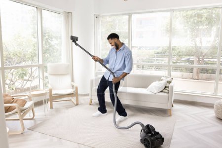 Photo for Cheerful Black homeowner guy having fun while tidying up at home, vacuuming carpeted floor in living room, dancing to music, singing song, imitating guitar playing with hoover - Royalty Free Image