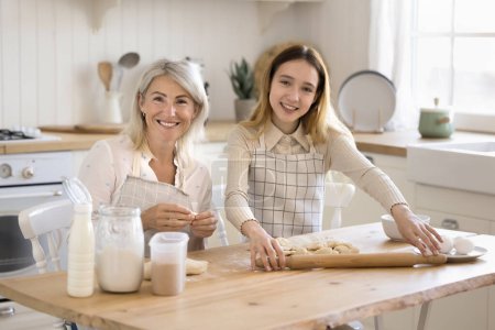 Beautiful middle-aged woman preparing pastries with cute teenager daughter in the kitchen, flattening homemade dough for holiday buns, teaching preteen girl to cook at home. Culinary, family recipe