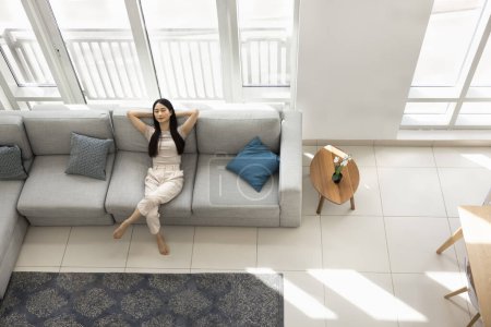 Téléchargez les photos : Belle jeune asiatique détendue reposant les yeux fermés sur un grand canapé confortable à la maison, profitant de la détente, du temps libre, respirant de l'air frais avec les coudes ouverts. Vue du dessus - en image libre de droit