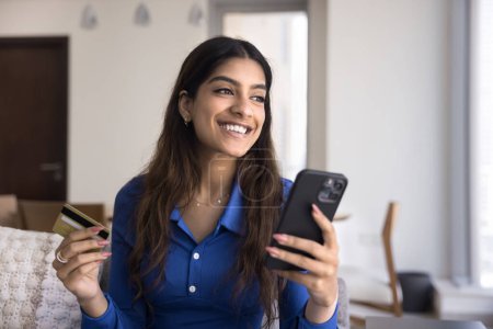 Foto de Joven consumidor indio alegre de ensueño disfrutando de compras en línea en el teléfono móvil, pagando con tarjeta de crédito, gastando dinero en compras en Internet, mirando hacia otro lado, pensando, sonriendo - Imagen libre de derechos