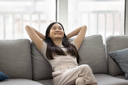 Photo for Cheerful relaxed beautiful Asian woman resting with closed eyes on comfortable couch at home, keeping hands on nap, smiling, breathing fresh pure air, enjoying homey comfort - Royalty Free Image