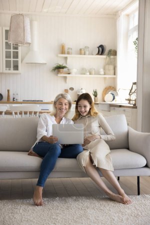 Photo for Happy teenager girl sit on sofa with middle aged mum in living room, watch funny videos in social media, browse internet, enjoy family weekend together. Two different ages generation use modern tech - Royalty Free Image