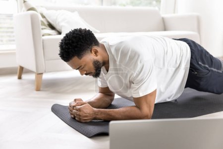 Photo for Focused strong sporty Black man training body at laptop, taking online fitness lesson, keeping forearm plank on mat, warming up. Athletic trainer giving class on Internet - Royalty Free Image