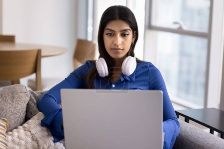 Photo for Serious young Indian student girl studying at home, holding laptop on lap, resting on home couch, typing, using big wireless headphones. Freelancer woman working on online communication project - Royalty Free Image