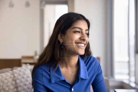 Téléchargez les photos : Joyeux jeune femme indienne attrayante maison portrait décontracté. Belle fille 20s positive regardant loin avec un sourire parfait, assise sur le canapé, rêvant, pensant avec des émotions positives - en image libre de droit