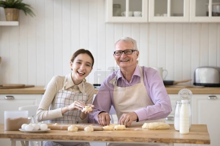 Téléchargez les photos : Happy preteen granddaughter and older grandpa wear aprons sit at table, cooking, prepare dessert, buns, sweets from homemade dough, smile, look at camera. Portrait of loving multigenerational family - en image libre de droit