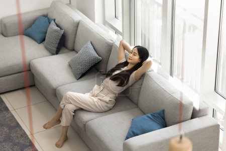 Calm beautiful Asian adult girl enjoying relaxation at cozy home, resting on comfortable spacious grey couch with hands on nap, taking deep breath of fresh air with closed eyes. High angle shot