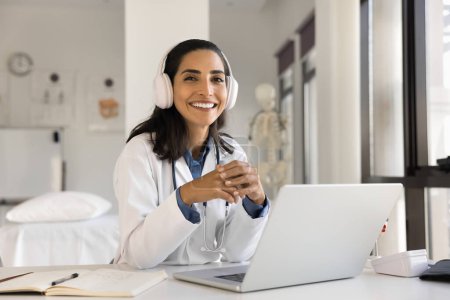 Photo for Cheerful beautiful young doctor woman in head phones posing at hospital workplace with laptop computer, looking at camera with toothy smile, offering online medical consultation. Professional portrait - Royalty Free Image