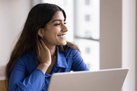 Photo for Happy Indian freelance worker girl sitting at laptop, touching neck in deep positive thoughts, looking away, smiling, dreaming, thinking on Internet communication technology - Royalty Free Image