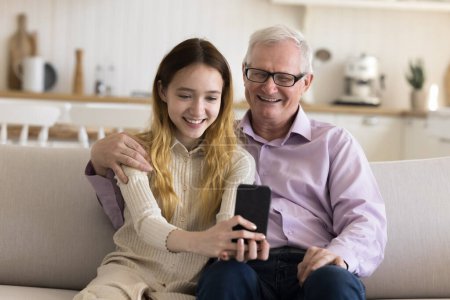 Téléchargez les photos : Pré-adolescente joyeuse prendre portrait de famille sur téléphone portable avec le vieux grand-père, en regardant l'écran de téléphone portable, sourire, faire une vidéoconférence avec la famille, profiter des loisirs insouciants à la maison. Technologie sans fil - en image libre de droit