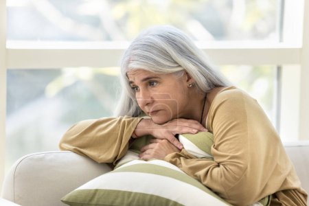 Photo for Depressed elderly woman sitting on home couch with sad pointless look away, embracing cushion, feeling bad, lonely, stressed, desperate, thinking on health problems - Royalty Free Image