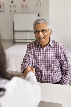 Foto de Agradecido positivo anciano indio paciente hombre dando apretón de manos a la doctora, terminando la reunión de examen de salud, hablando, sonriendo, riendo, agradeciendo por la consulta, terapia eficaz - Imagen libre de derechos