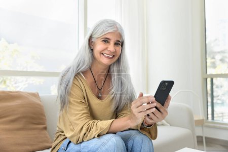 Téléchargez les photos : Enthousiaste femme latine âgée aux cheveux gris positifs utilisant une application en ligne, service de commerce électronique sur téléphone mobile, posant pour portrait à la maison sur canapé blanc, regardant la caméra avec le sourire aux dents - en image libre de droit