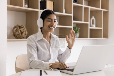Photo for Cheerful young Indian manager woman in headphones online meeting, job communication on Internet, talking on video conference call, waving greeting hand hello at laptop - Royalty Free Image