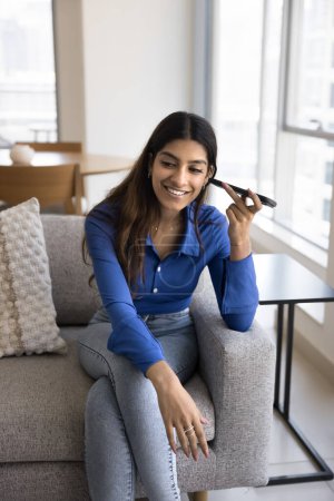 Photo for Cheerful young Indian woman listening to voice message from online chat, sitting on comfortable couch at home, using mobile phone, holding dynamic at ear, smiling. Vertical shot - Royalty Free Image