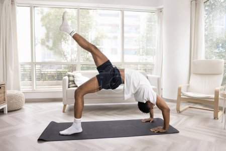 Photo for Strong athletic young African man doing bridge stand with raising leg at home, keeping wheel backbend pose on yoga mat, stretching front muscles, exercising in morning - Royalty Free Image