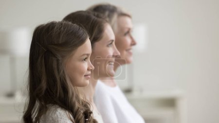 Photo for Side profile faces view of happy three generation of relatives women. Beautiful preschooler girl looking forward standing posing indoors in row with young pretty mother and senior cheerful grandmother - Royalty Free Image
