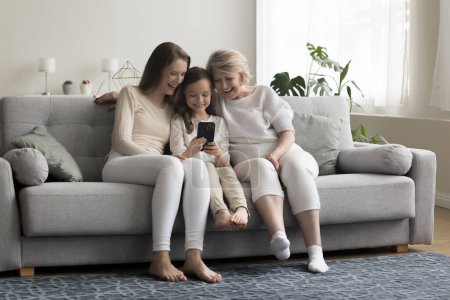 Mehrgenerationenfamilien verbringen ihre Freizeit mit der Internetnutzung von Mobiltelefonen, sehen Videos an und lachen, genießen neue mobile amüsante Applikationen, sitzen zusammen auf dem Sofa im Wohnzimmer. Moderne Technik, Spaß, Zeitvertreib