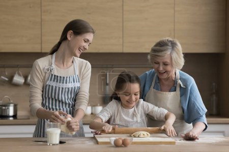 Photo for Little girl her grandmother and loving young mom wear aprons cooking together in kitchen, flattening prepared dough making homemade delicious buns, enjoy communication, pastime together and cookery - Royalty Free Image