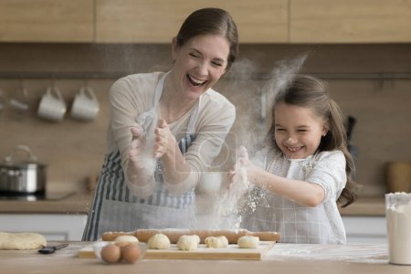 Photo for Happy young mother and little 6s cute daughter having fun while cooking pastries in domestic kitchen, clap their hands sprinkling white flour, laughing. Family fooling around during food preparation - Royalty Free Image