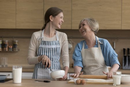 Photo for Laughing millennial woman in apron whisking mixture for cake, cooking together with cheerful aged mother holding rolling-pin, flattening dough for homemade pastries, spend weekend in modern kitchen - Royalty Free Image
