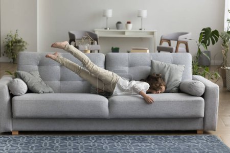 Photo for Adorable little girl enjoy carefree pastime at modern home, jump on cozy couch in living room, entertain on comfortable furniture, looks happy take pleasure on weekend. Exhaustion relief, relaxation - Royalty Free Image