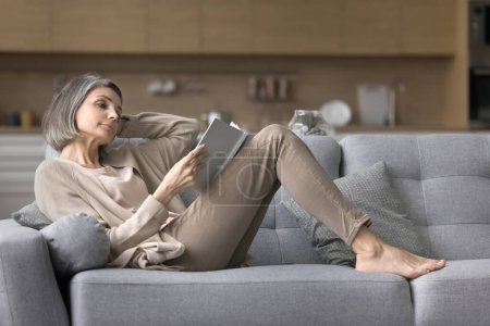 Photo for Pretty mature woman resting on comfortable sofa with book, feel satisfied reading interesting literature, favorite novel or poetry, spend weekend time alone indoors, booklover, hobby, pastime at home - Royalty Free Image