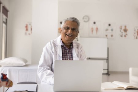 Photo for Happy mature senior Indian doctor man wearing white uniform and glasses, sitting at clinic workplace table, working at laptop, looking at camera, laughing, smiling, posing for portrait - Royalty Free Image