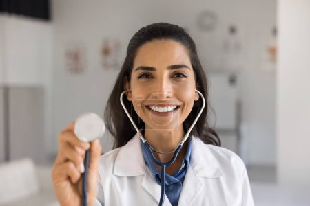 Photo for Happy young Latin doctor using stethoscope head shot portrait. Positive practitioner woman, cardiologist, therapist promoting healthcare examination, showing work tool at camera, smiling - Royalty Free Image