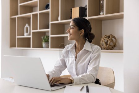 Photo for Cheerful successful young Indian manager woman using laptop in office, looking away, thinking on creative ideas for business project, enjoying professional inspiration, job success - Royalty Free Image