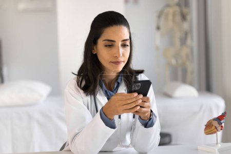 Téléchargez les photos : Jeune cardiologue latine sérieuse vérifiant les patients, tapant des messages sur un téléphone portable, bavardant en ligne, utilisant un smartphone pour garder la connexion, donnant une consultation en ligne - en image libre de droit